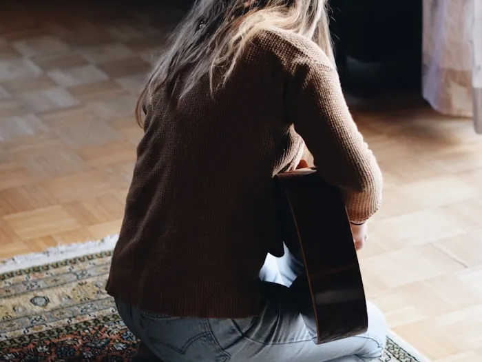 A woman kneeling on a rug, playing a guitar in a warm home interior